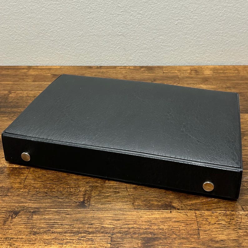 May include: A black, rectangular binder with a textured surface. The binder has two silver-colored metal rivets on the spine. The binder is resting on a wooden surface.