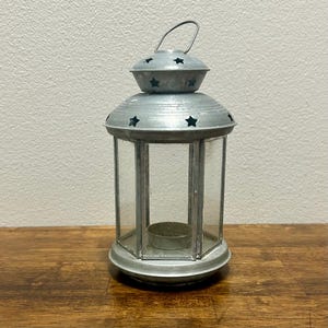 May include: A silver-colored metal lantern featuring a hexagonal glass body and a tiered roof. The roof and the smaller top cap are decorated with cut-out star shapes. A small metal handle is attached to the top, and a circular candle holder sits inside.