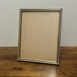 May include: A rectangular picture frame with a metallic gold finish and a textured, beaded edge detail. The frame contains a plain, light brown cardboard insert that fills the entire interior space, providing a neutral display area.
