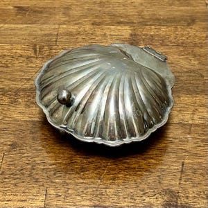 May include: A silver-colored, shell-shaped trinket box with a hinged lid and a small, round knob. The box has a detailed, ridged design and sits on a wooden surface.
