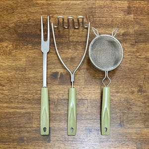 May include: A set of three vintage kitchen utensils: a fork, a potato masher, and a strainer. Each tool has a silver metal head and a light green plastic handle. The utensils are arranged on a wooden surface.