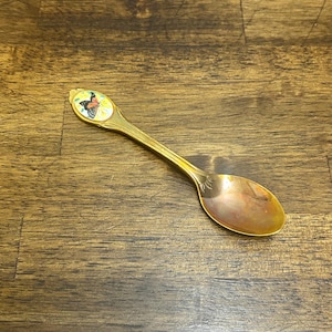 Può includere: Un cucchiaio color oro con un disegno a farfalla sul manico. La farfalla è raffigurata con dettagli rossi, gialli e neri. Il cucchiaio ha una forma arrotondata ed è appoggiato su una superficie di legno.