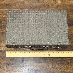 May include: A rectangular, vintage case with a patterned, olive green and pink fabric exterior. The case has a silver-colored handle and clasps. A wooden ruler is placed below the case.