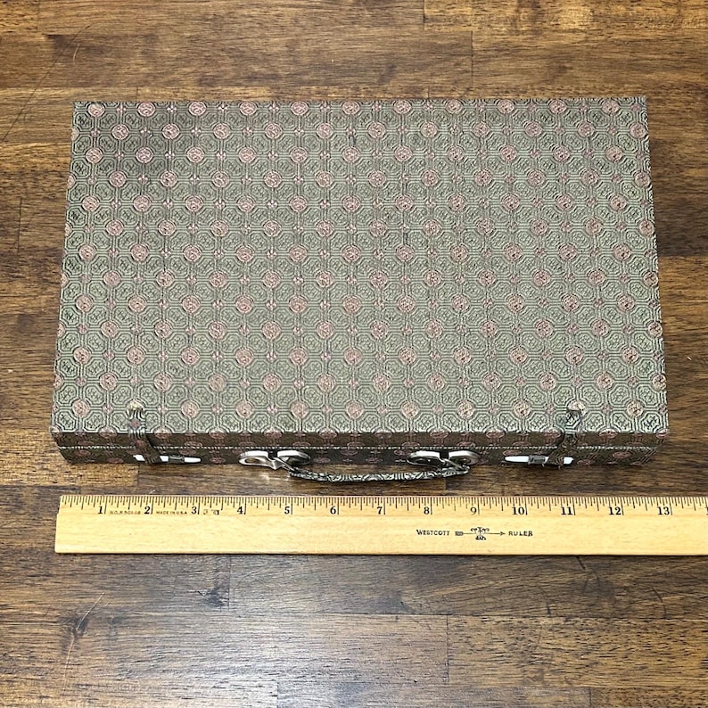 May include: A rectangular, vintage case with a patterned, olive green and pink fabric exterior. The case has a silver-colored handle and clasps. A wooden ruler is placed below the case.
