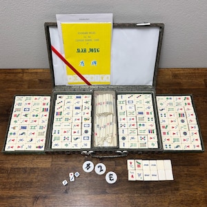 May include: An open Mah Jong set with tiles, dice, and counters on a wooden surface. The set includes a case with multiple compartments, a yellow booklet with the title "STANDARD RULES for the CHINESE DOMINO GAME of MAH JONG", and a red ribbon.