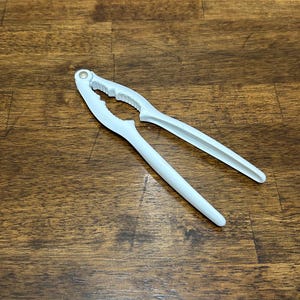 Puede incluir: Un cascanueces blanco de plástico, sobre una superficie de madera. La herramienta está abierta, mostrando el mecanismo de agarre. Está diseñado para romper nueces.