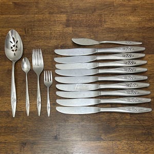 May include: A set of vintage silverware, including knives, forks, spoons, and a serving spoon. The silverware features a floral design on the handles and is arranged on a wooden surface. The set includes 12 knives.