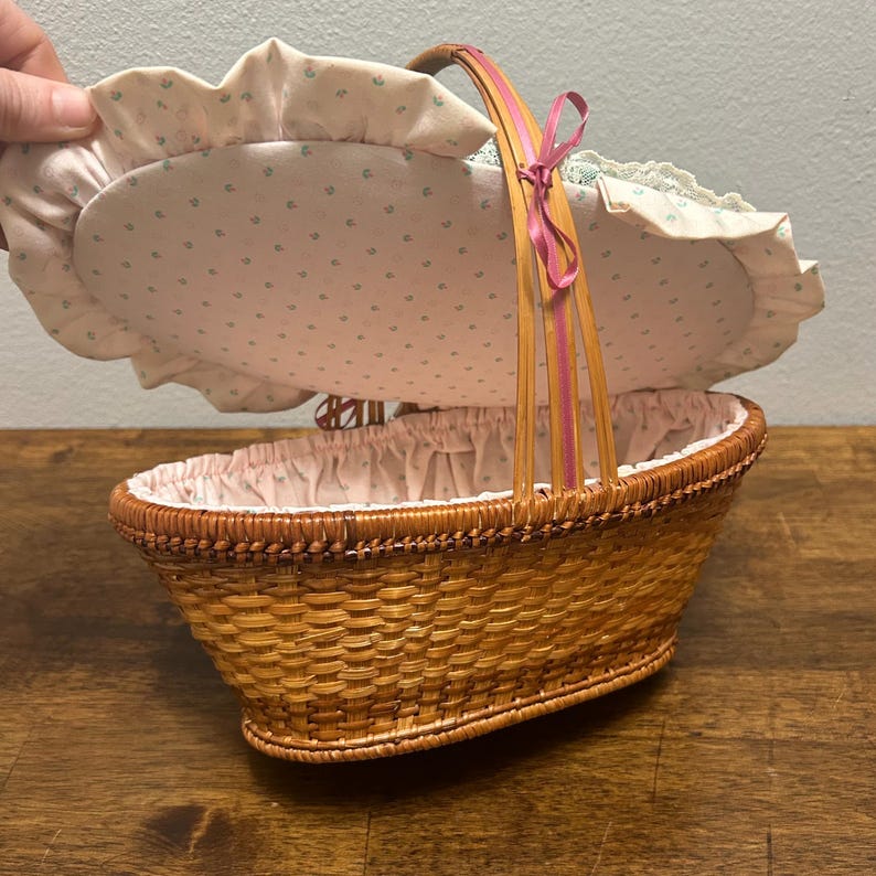 Puede incluir: Una cesta ovalada de mimbre trenzado con tapa abatible que presenta un forro de tela rosa claro con un peque&ntilde;o estampado floral verde azulado y un borde con volantes. La cesta tiene un asa arqueada alta con una cinta rosa fina en la parte superior.