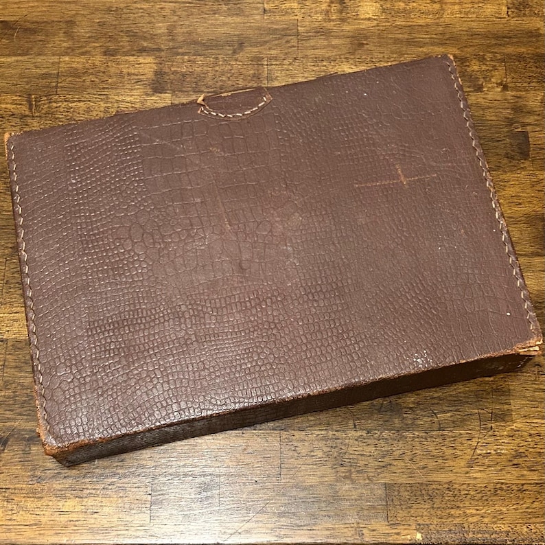May include: A rectangular brown storage box with a textured faux-alligator skin pattern on the exterior. The box features visible stitching along the edges and a small, semi-circular tab closure at the top center of the lid, showing signs of wear.