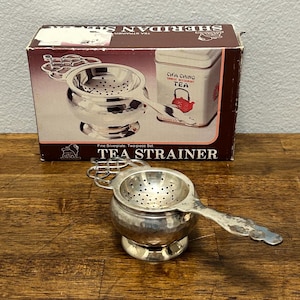 May include: A silver-colored tea strainer with a handle, sitting in front of a box. The box reads "TEA STRAINER" and "CHA CHING CHINESE RESTAURANT TEA". The strainer has a bowl shape with small holes for filtering tea leaves.
