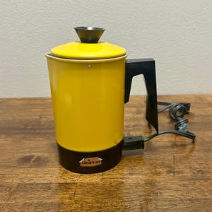 May include: A bright yellow electric percolator with a black base, handle, and lid knob. The appliance features a small rectangular label on the front with the word Chilton printed in black text. A black power cord is attached to the side.