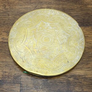 May include: A circular gold-toned metal compact mirror featuring an intricate engraved pattern on the lid. The design consists of a central star-like motif surrounded by detailed scrollwork, floral elements, and geometric lines across the surface.