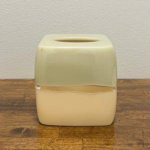 May include: A two-tone tissue box in a square shape. The top half is a light green color, and the bottom half is a light beige color. The tissue box is sitting on a wooden surface.