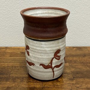 May include: A cylindrical ceramic vessel featuring a textured off-white body with hand-painted reddish-brown floral motifs. The upper rim is finished in a smooth, solid dark brown glaze, separated from the lower section by a thin black line.