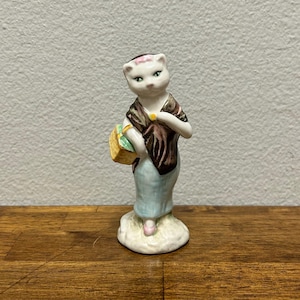 May include: A ceramic figurine of a cat-like figure, wearing a shawl and a light blue dress, holding a basket. The figurine has a white face with green eyes and a pink bow. The shawl is brown and the basket is yellow and green.