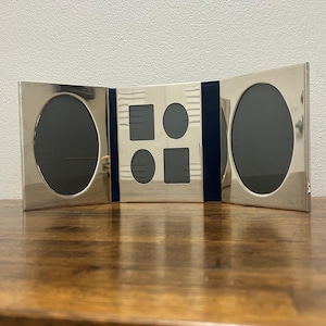 May include: A silver-toned triptych photo frame featuring two large oval openings on the side panels and a central panel containing two smaller rectangular openings and two smaller oval openings, all connected by dark blue fabric hinges.