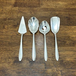 May include: Four silver serving utensils are displayed on a wooden surface. The set includes a pie server, a serving spoon with a scalloped bowl, a slotted spoon, and a serving spoon with a square bowl. The utensils are reflective and appear to be vintage.