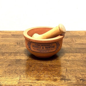May include: A terracotta pestle and mortar set. The mortar is a round bowl with a wide rim, and the pestle is a wooden, cylindrical shape. The set has a label that reads "PESTLE & MORTAR" and "Made by Henry Watson Pottery England Founded 1800".