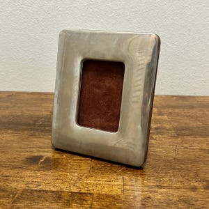 May include: A rectangular picture frame with a polished silver-toned metallic border and a dark brown velvet-textured insert. The frame features a smooth, reflective surface and a simple, clean design with rounded corners on the outer edge.