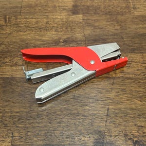May include: A red and silver stapler, a common office supply, is shown. The stapler has a red handle and a silver metal body. The stapler is designed to hold and dispense staples for fastening papers together. The stapler is on a wooden surface.