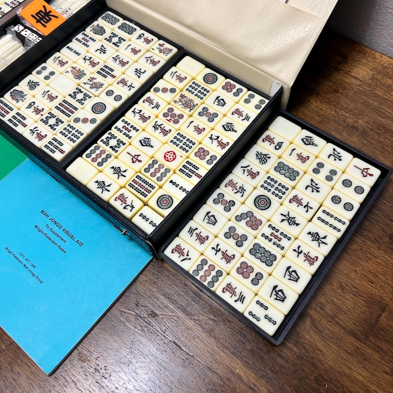 May include: A vintage Mahjong set with ivory-colored tiles, each marked with Chinese characters and symbols, neatly arranged in black trays within a cream-colored case. A blue booklet with the text "Mah Jongg Visual Aid" is visible.