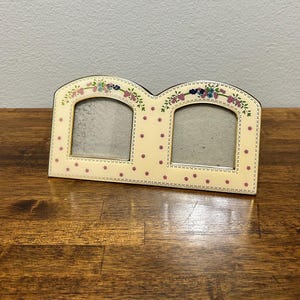 Puede incluir: Un marco de fotos doble con fondo crema y detalles florales. El marco tiene dos aberturas arqueadas y está decorado con flores rosas y hojas verdes. El marco está sobre una superficie de madera.