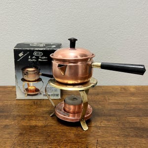 May include: A copper butter warmer set with a small saucepan, lid, and stand. The saucepan has a black handle and a spout. The stand is brass-colored with a place for a tea light. A box with the product name is in the background.