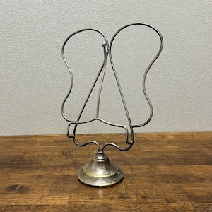 May include: A silver-toned metal display stand featuring a curved, open-wire frame shaped like butterfly wings. The stand has a sturdy, circular base with a tarnished metallic finish and a central support arm designed to hold items upright.