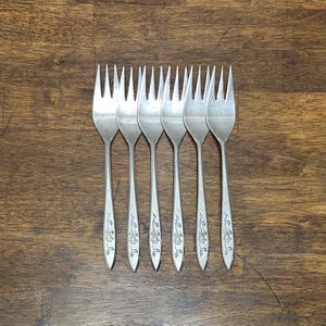 May include: A collection of six silver forks, each featuring a floral pattern on the handle. The forks are laid out on a wooden surface. The tines are aligned, and the handles have a delicate design. This cutlery set is suitable for a formal dinner.