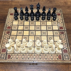 May include: A folding wooden chessboard featuring an intricate inlaid geometric border pattern in shades of brown, cream, and black. The board is set with a complete set of black and cream-colored plastic chess pieces, including kings, queens, bishops, knights, rooks, and pawns, arranged in their starting positions on the board.