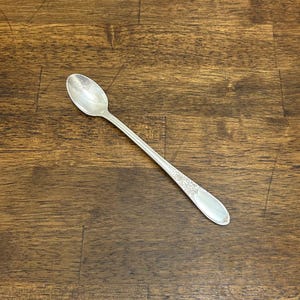 Puede incluir: Una cuchara plateada con un mango largo y un cuenco ovalado. El mango presenta un diseño floral ornamentado cerca del extremo del cuenco. La cuchara está sobre una superficie de vetas de madera oscura.