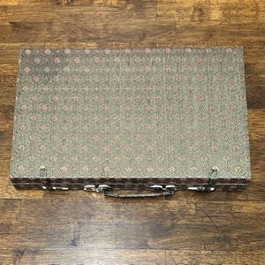 May include: A rectangular, closed case with a patterned fabric exterior. The fabric has a repeating design in shades of green and pink. The case has a silver-colored handle and clasps.