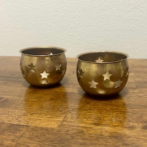 May include: Two small, round, metallic candle holders with a brass-toned finish. Each bowl-shaped vessel features a series of cut-out star shapes around the exterior circumference, allowing light to pass through the openings in the metal walls.