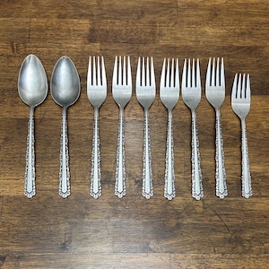 May include: A collection of silverware, including two spoons and seven forks, arranged on a wooden surface. The silverware has ornate, detailed handles. The spoons and forks are made of a shiny, silver-colored metal.