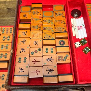 May include: A vintage Mahjong set featuring orange tiles with black, green, and red engraved characters, symbols, and illustrations. The set includes white counting sticks, two green translucent dice, and one white die, all housed in a red felt-lined case.