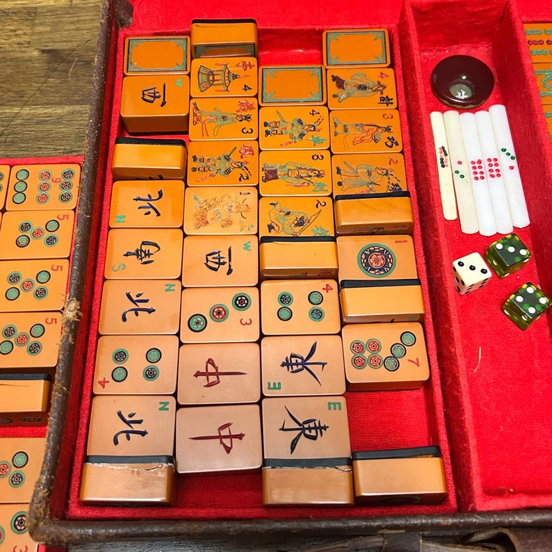 May include: A vintage Mahjong set featuring orange tiles with black, green, and red engraved characters, symbols, and illustrations. The set includes white counting sticks, two green translucent dice, and one white die, all housed in a red felt-lined case.
