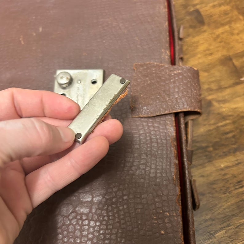 May include: A brown textured suitcase with a detached silver metal latch piece held in the foreground. The suitcase features a rectangular silver locking mechanism mounted on the front, with a matching leather strap attached to the side edge.