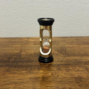 May include: A small hourglass with a gold-colored metal frame and black top and bottom. The glass chamber contains brown sand. The hourglass is a decorative item, likely used for timing or as an ornamental piece.