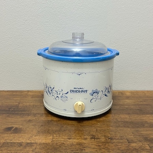 以下が含まれることがあります： 青い縁と蓋が付いた、白いRival Crock-Potストーンウェアスロークッカー。スロークッカーは青い花柄のデザインで、「RIVAL CROCK-POT」の文字が入っています。コントロールノブには、オフ、ロー、ハイの設定があります。