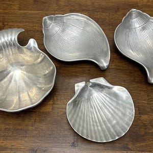 May include: Four silver-colored seashell-shaped dishes on a wooden surface. The dishes vary in design, with some having a textured, ridged appearance and others featuring a smooth, curved form. The arrangement suggests a coastal or nautical theme.