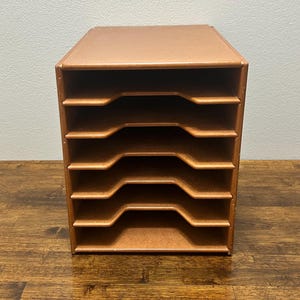 May include: A brown wooden desk organizer with six open compartments. The organizer is rectangular and has a smooth finish. The compartments have a curved design for easy access. The organizer is designed for office or home use.