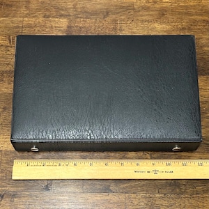 May include: A rectangular black case with a textured surface, approximately 12 inches long. Two silver-colored metal clasps are visible on the front. A wooden ruler is placed below the case for scale.