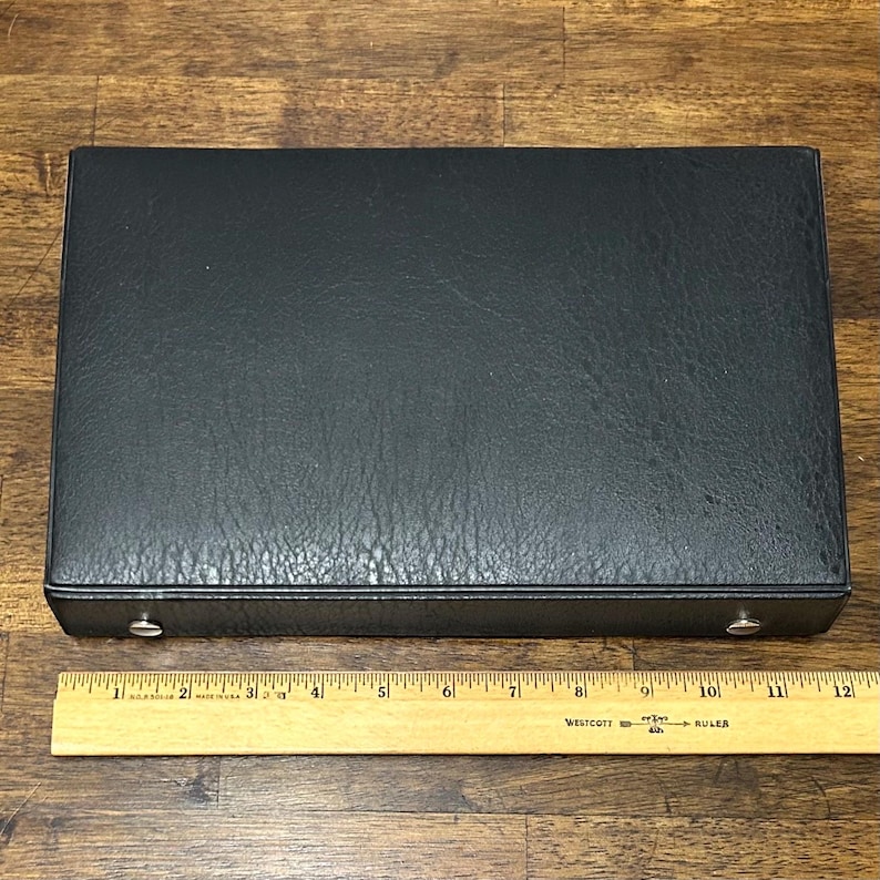 May include: A rectangular black case with a textured surface, approximately 12 inches long. Two silver-colored metal clasps are visible on the front. A wooden ruler is placed below the case for scale.