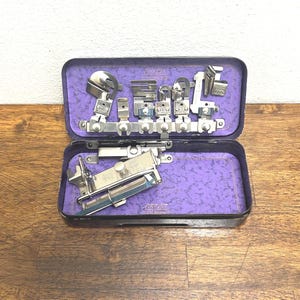 May include: A rectangular black metal case with a purple interior containing various silver-colored metal sewing machine attachments. The case features the text Greist Attachments printed in white on the interior lid and base.