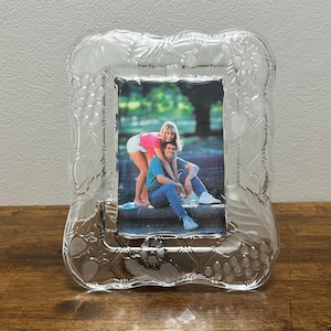 May include: A rectangular clear glass picture frame featuring a decorative border with etched patterns of grapes, leaves, and flowers. The frame holds a photograph of two people sitting together outdoors in a park setting with green trees.