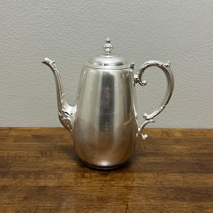 May include: A silver-colored coffee pot with a curved spout and ornate handle. The pot has a lid with a decorative finial and sits on a wooden surface. The pot's surface reflects the surrounding environment.