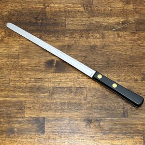Puede incluir: Un cuchillo largo y dentado de acero inoxidable con un mango negro y dos remaches dorados. El cuchillo est&aacute; dise&ntilde;ado para cortar y se encuentra sobre una superficie de vetas de madera oscura.