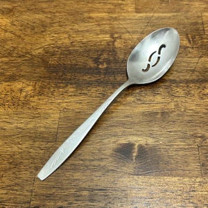 May include: A silver slotted spoon with a floral design on the handle. The spoon has two curved slots in the bowl. The spoon is made of stainless steel and is designed for serving food.
