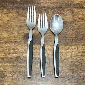 May include: A stainless steel flatware set featuring two forks and a spoon. The utensils have black handles with a decorative silver design near the top. The set is displayed on a wooden surface.