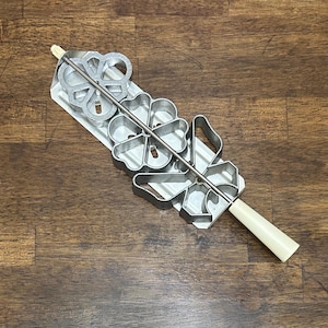 Puede incluir: Una plancha de rosetas de metal vintage con varios cortadores de galletas con forma de flor. La plancha tiene un mango largo de color blanquecino y una varilla de metal que sujeta los cortadores en su lugar. La plancha de rosetas está diseñada para hacer galletas decorativas.